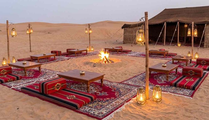 A traditional Bedouin-style desert camp setup at dusk, featuring low wooden tables, vibrant red rugs, and cushions arranged around a central campfire. Numerous hanging lanterns cast a warm glow over the scene, which is set amongst sand dunes with a large tent in the background, providing a perfect setting for an Anniversary Dinner In Abu Dhabi Desert