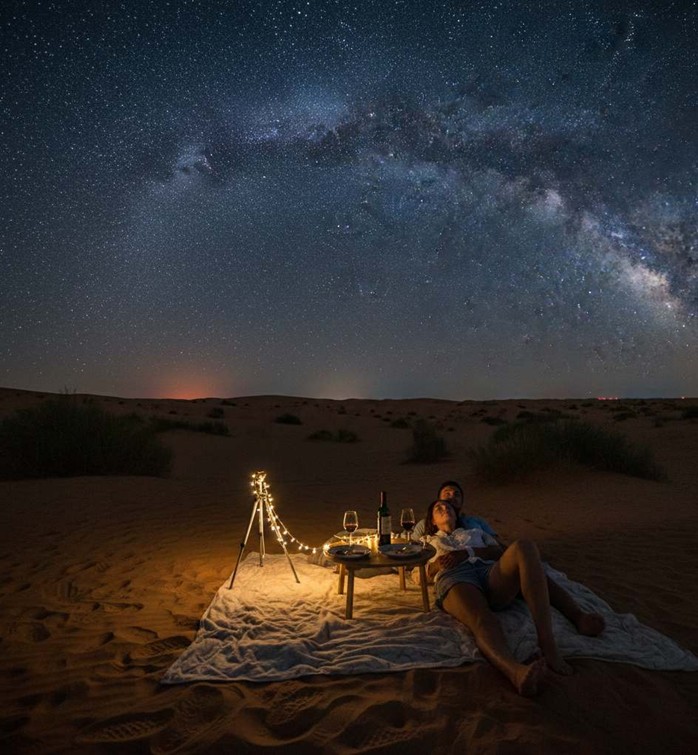 A couple relaxes on a blanket after an Anniversary Dinner In Abu Dhabi Desert, lying back to gaze at a brilliant Milky Way galaxy sprawling across the clear, dark night sky above the rolling sand dunes.