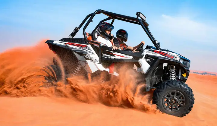Two helmeted riders in a white Polaris dune buggy speeding across red sand dunes in the Abu Dhabi Desert, kicking up a large cloud of sand.