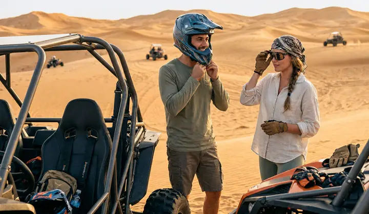 Man and woman preparing for a dune buggy ride in the Abu Dhabi Desert; he adjusts his blue helmet while she adjusts her sunglasses, with buggies and sand dunes behind them.