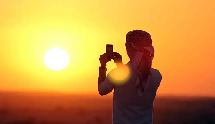 Silhouette of a man wearing a traditional Ghutra taking a photo of the golden rising sun using his smartphone.