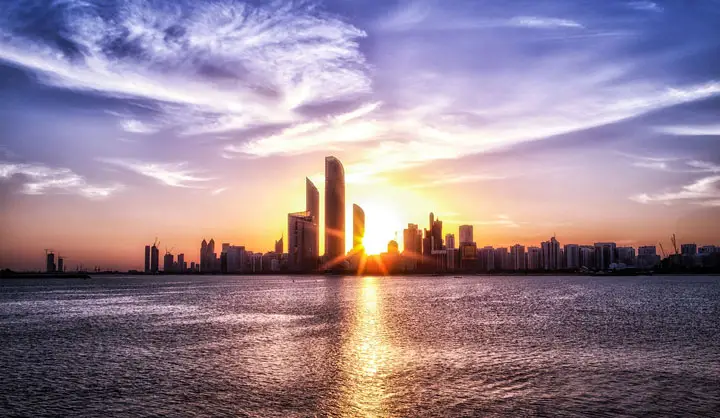Sun rising directly behind the Abu Dhabi city skyline illuminating the calm coastal waters.