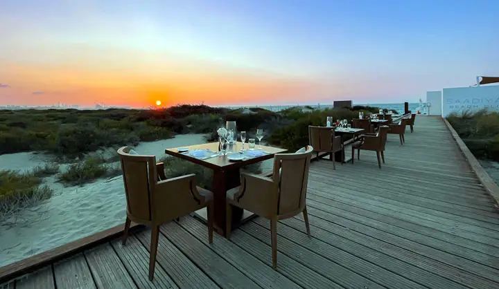 Outdoor dining tables set up on a wooden deck at Saadiyat Beach Club overlooking the beach at sunrise in Abu Dhabi.