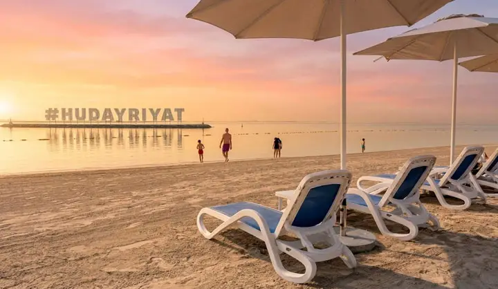 Empty beach lounge chairs lined up on Hudayriyat Beach with the Hudayriyat sign in the background at sunrise.