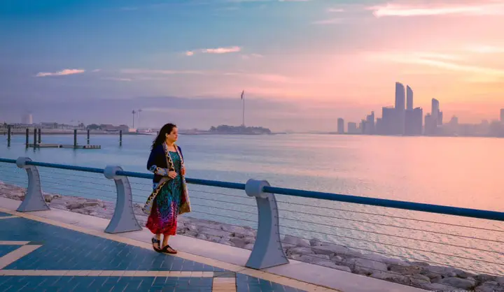 Woman walking along the Corniche promenade with the Abu Dhabi skyline reflecting off the water in soft morning light.