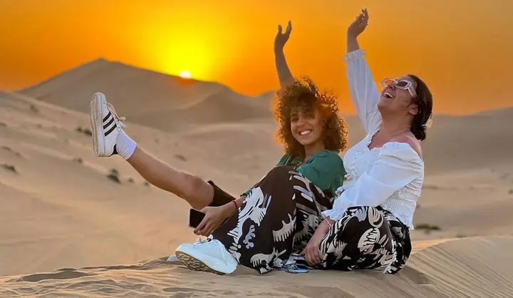 Two joyful women sitting on a sand dune cheering with arms raised at sunrise in the Abu Dhabi desert.
