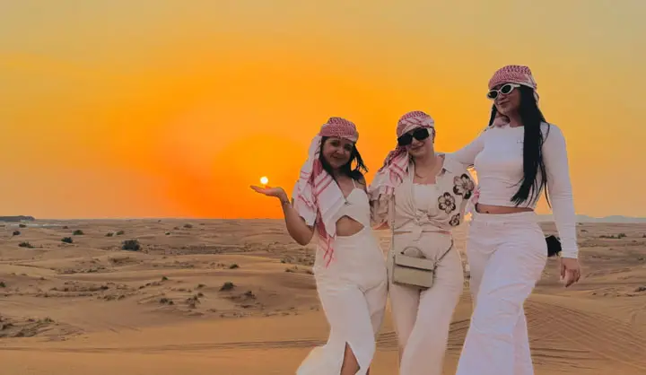 Three women wearing traditional headscarves posing against a glowing sun over sand dunes during a sunrise safari in Abu Dhabi.