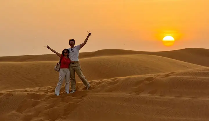 Happy couple standing on desert dunes waving their arms as the bright sun rises over the horizon in Abu Dhabi.