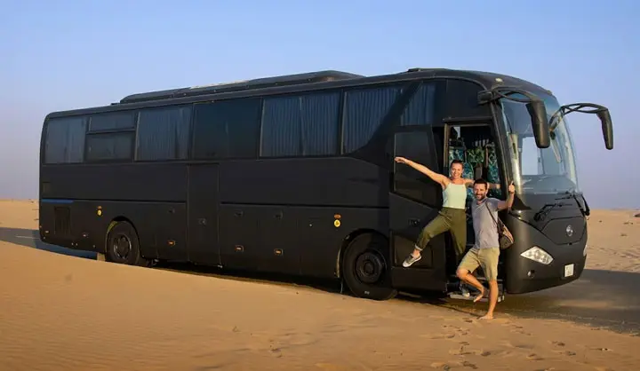 Happy couple posing enthusiastically at the open doorway of a large black tour bus parked in the Abu Dhabi desert sand.