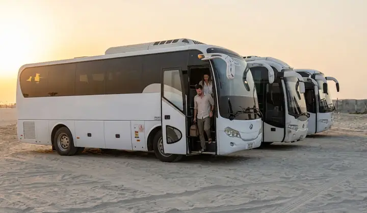 Tourists stepping down from a white luxury coach bus lined up with a fleet of buses in the Abu Dhabi desert.