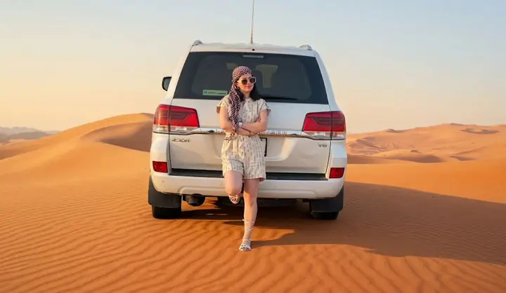 Stylish woman wearing a headscarf leaning against a white Toyota Land Cruiser SUV on the golden sand dunes of Abu Dhabi.