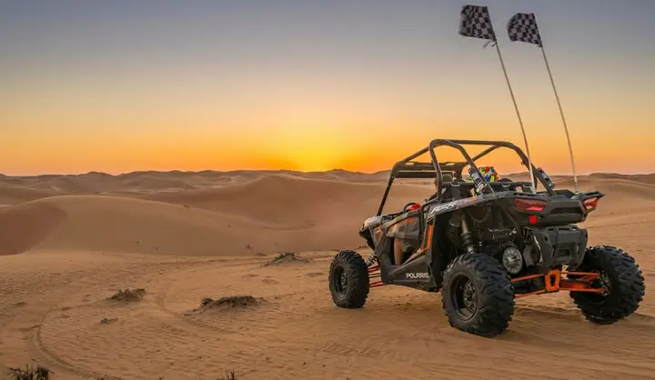 Black Polaris dune buggy parked on golden sand dunes in Abu Dhabi desert at sunset, with two checkered flags waving behind it.