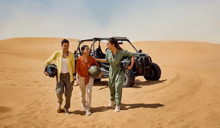 Three smiling women walking across Abu Dhabi desert sand dunes carrying helmets, with a dune buggy parked behind them.