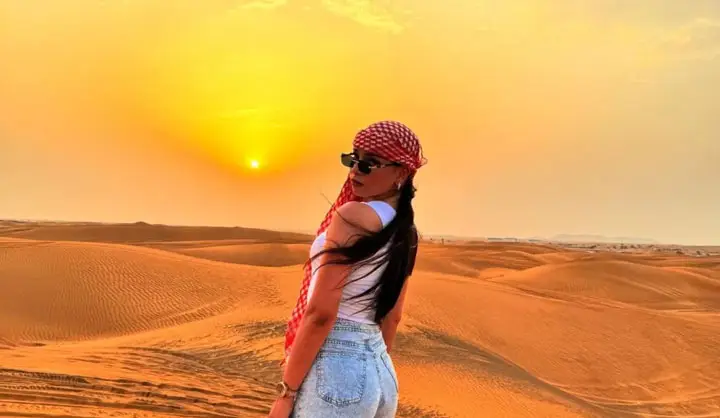 Stylish woman wearing a traditional keffiyeh headscarf and sunglasses posing against a glowing sunset in the Abu Dhabi desert.