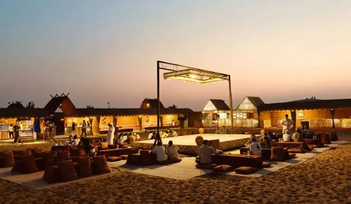 Bedouin-style evening desert camp in Abu Dhabi featuring low seating cushions around a lit central stage at dusk.
