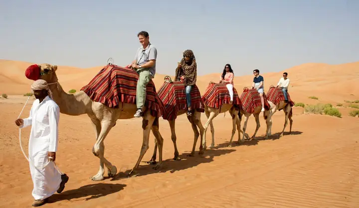 A group of tourists enjoying a camel caravan trek across the vast orange dunes of the Abu Dhabi desert, led by a guide in traditional white attire.