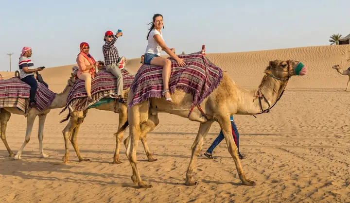 Happy tourists riding decorated camels through the Abu Dhabi desert, with one woman smiling at the camera while others take photos during the trek.