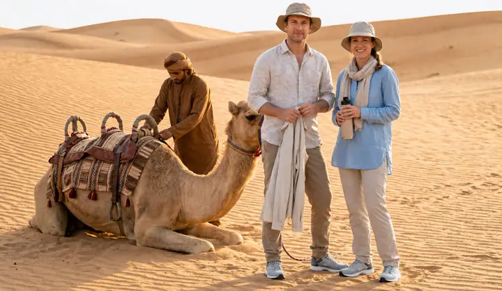Tourists pose beside a resting camel in the Abu Dhabi desert while a local handler adjusts its saddle.