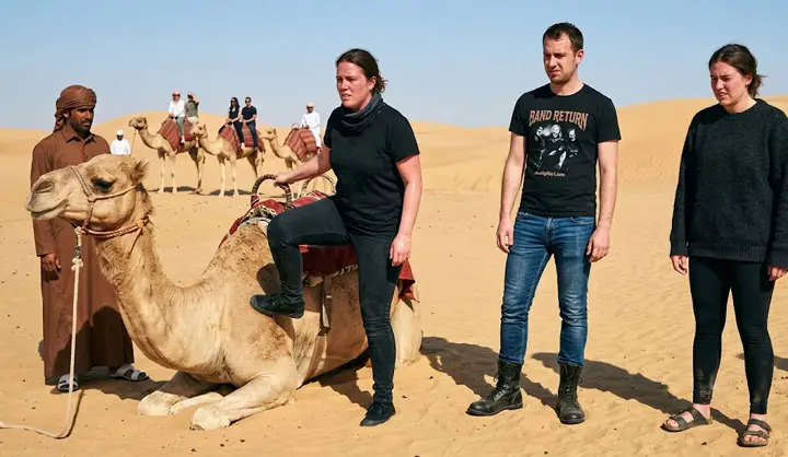 Tourists stand beside a resting camel in the Abu Dhabi desert, one woman seated on its saddle.