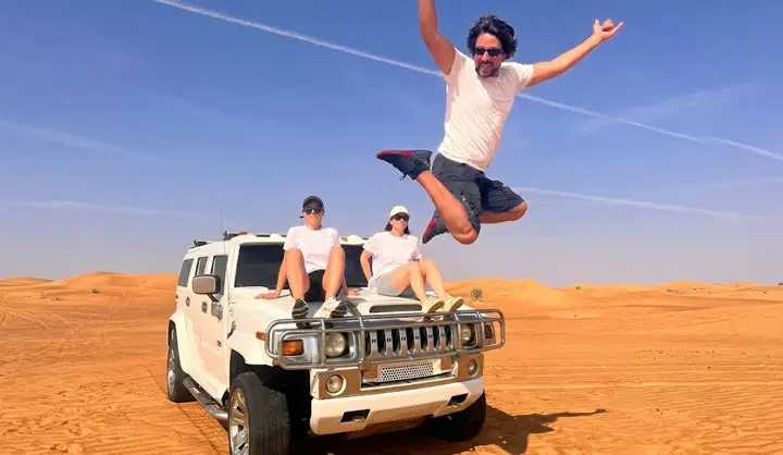 Group of friends wearing casual summer clothes posing and jumping in front of a white Hummer SUV in the Abu Dhabi desert.