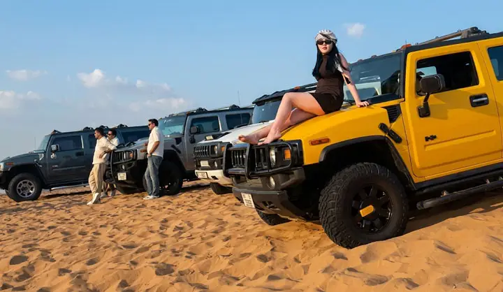 Woman sitting on the hood of a bright yellow Hummer SUV with a fleet of off-road vehicles in the Abu Dhabi desert.