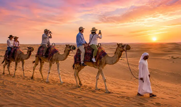 A line of tourists riding camels through sandy desert dunes at sunset, led by a guide wearing traditional Emirati attire under a vibrant pink and orange sky.