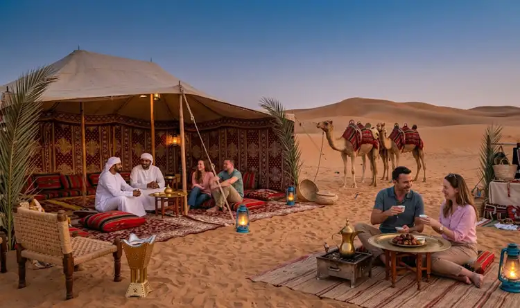 Tourists and local hosts relaxing, drinking coffee, and eating dates outside a traditional patterned Bedouin tent in the desert at dusk, with camels standing nearby.