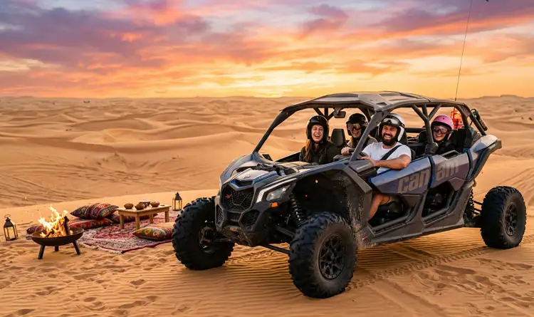 Four cheerful family members wearing helmets inside a dark blue Can-Am buggy parked on a sand dune next to a lit fire pit and a traditional floor seating arrangement.