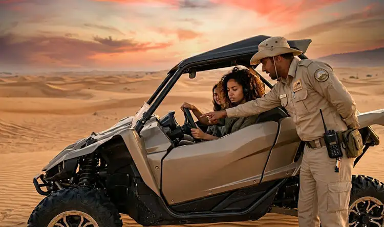 A desert safari guide in uniform pointing ahead to give driving directions to two women sitting in a tan off-road vehicle amidst the sand dunes.